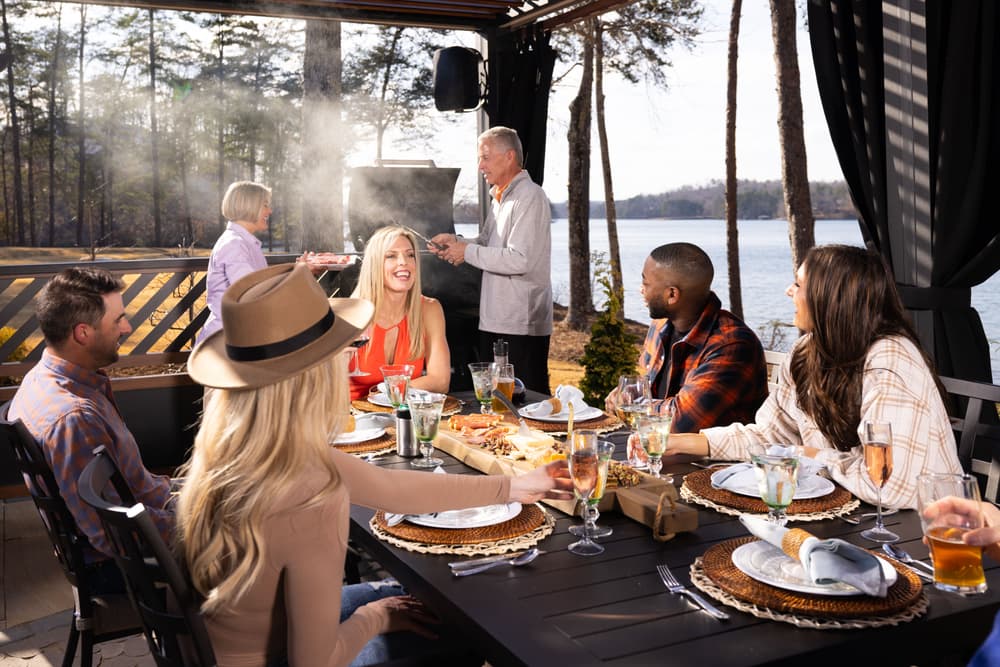 Outdoor barbecue gathering by the lake with friends enjoying food and drinks.